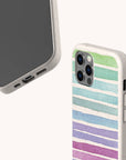 The Winter Rainbow Eco-Friendly Phone Case in speckled beige features pastel horizontal stripes in green, blue, purple, and pink. The smartphone is displayed from the back and bottom angles against a plain light background.
