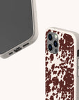 Two smartphones with beige cases—Brown Cow Print Eco-Friendly Phone Case designs; one showcases a brown cow print splatter pattern, while the other remains plain. Compatible as iPhone and Samsung Galaxy case options, both feature precise cutouts for ports and camera. The phones are angled to display both front and back views.