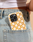 A Star Terracotta Checker Eco-Friendly Phone Case with a yellow and white checkered floral design sticks out of the back pocket of blue jeans, worn by a person in a white crop top.