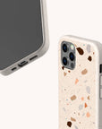 Two smartphones in beige Terrazzo Print Eco-Friendly Phone Cases with brown, tan, and orange flecks—one shows the back and camera, the other the screen and port. Sustainable protection meets modern style.