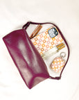 A plum leather pouch on a cream surface sits partially open, revealing a Star Pink Checker Eco-Friendly Phone Case, matching AirPods case, perfume bottle, and two gold makeup items inside.