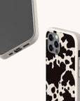 Two smartphones in beige cases—one features the Cow Print Eco-Friendly Phone Case, while the other is plain. Both eco-friendly cases fit iPhone and Samsung Galaxy, shown from different angles on a light background.