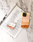 A smartphone in a vibrant Mountains Eco-Friendly Phone Case, plus wireless earbuds and their open pink case, sit on a rolled magazine atop marble. Compatible with iPhone and Samsung Galaxy—ideal for sustainable design lovers.