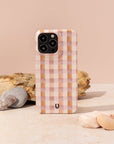 A Pink and Brown Gingham Eco-Friendly Phone Case stands upright on a light surface amid smooth stones and a textured rock, set against a soft peach background.