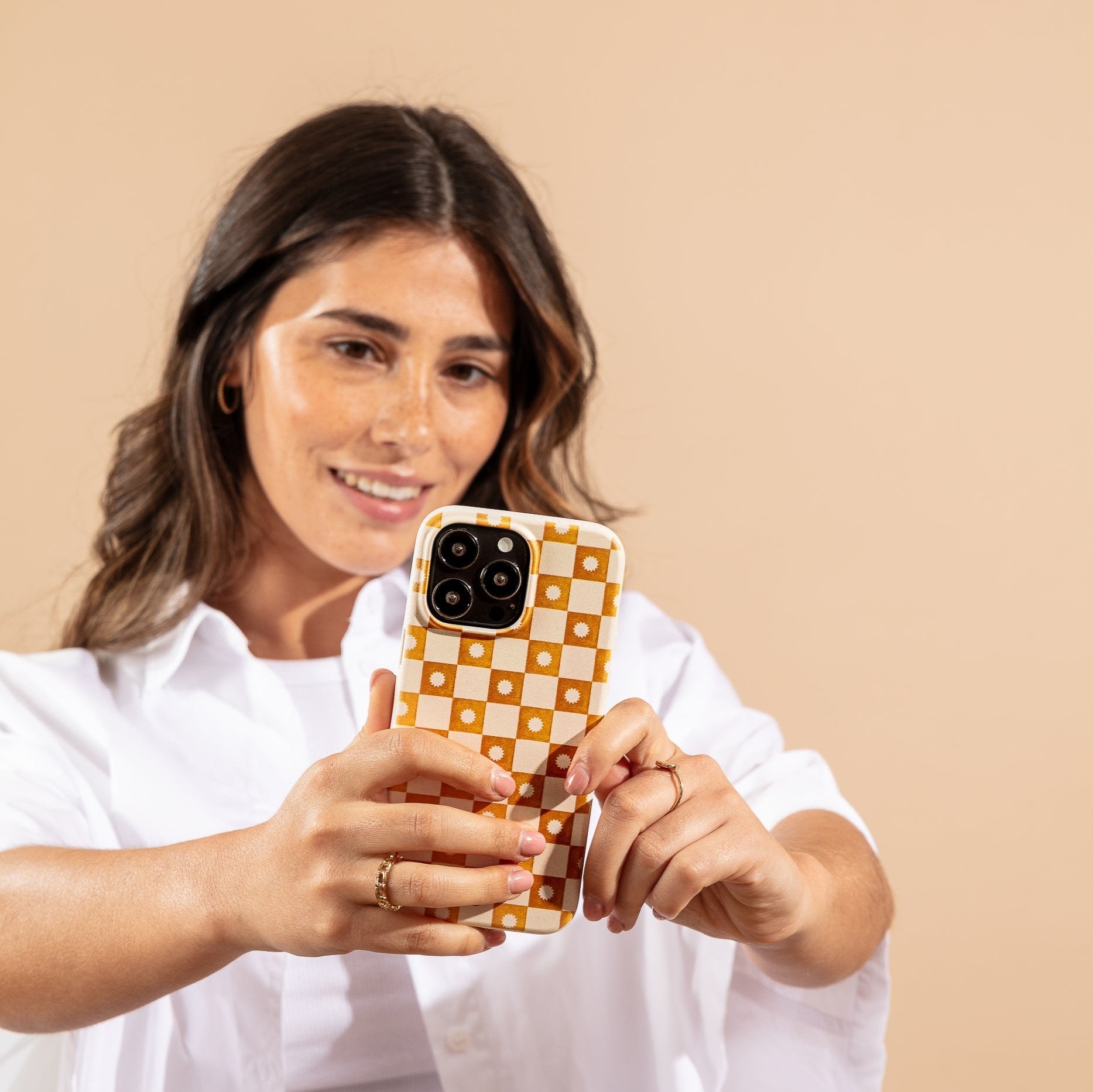 A woman with long brown hair and a white shirt smiles as she takes a selfie against a beige background, holding her smartphone with the Star Terracotta Checker Eco-Friendly Phone Case in yellow and white checks.