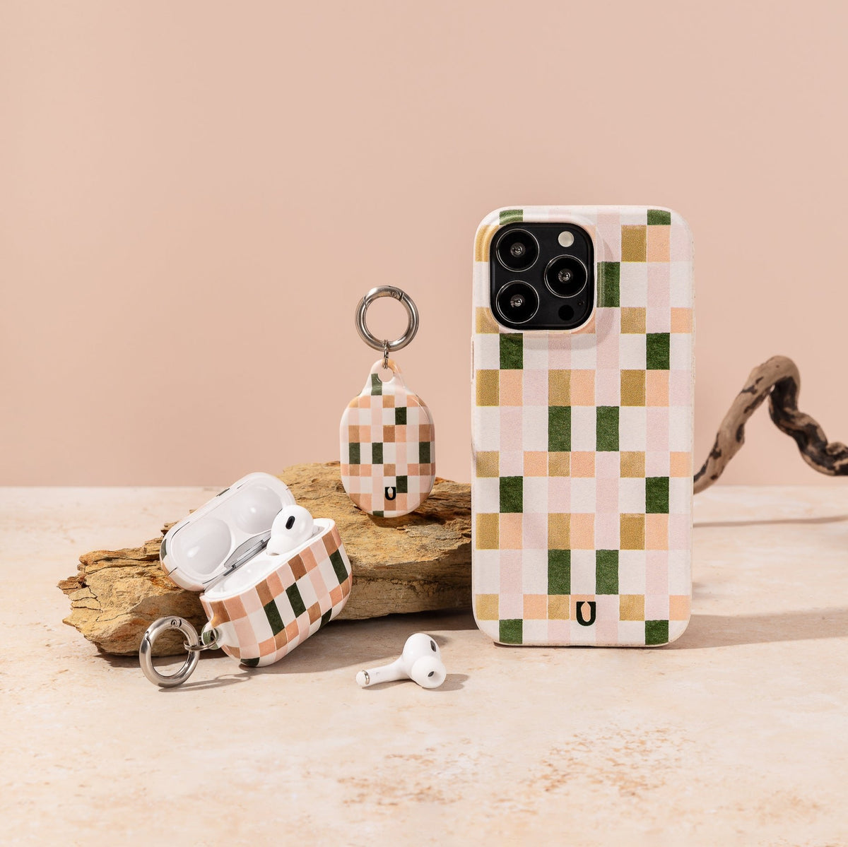 A phone with the Pink and Green Watercolour Eco-Friendly Phone Case, along with AirPods in a matching case and a keychain accessory—all featuring a pastel green, pink, and cream checkered pattern—are displayed on a beige surface with a piece of wood in the background.
