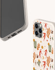 Two smartphones with beige cases—one displaying the front, the other highlighting the back featuring the vibrant “Three Dancers Eco-Friendly Phone Case” adorned with red figures, orange hands, green plants, and small yellow suns against a speckled background. These eco-friendly phone cases combine striking design with sustainability.