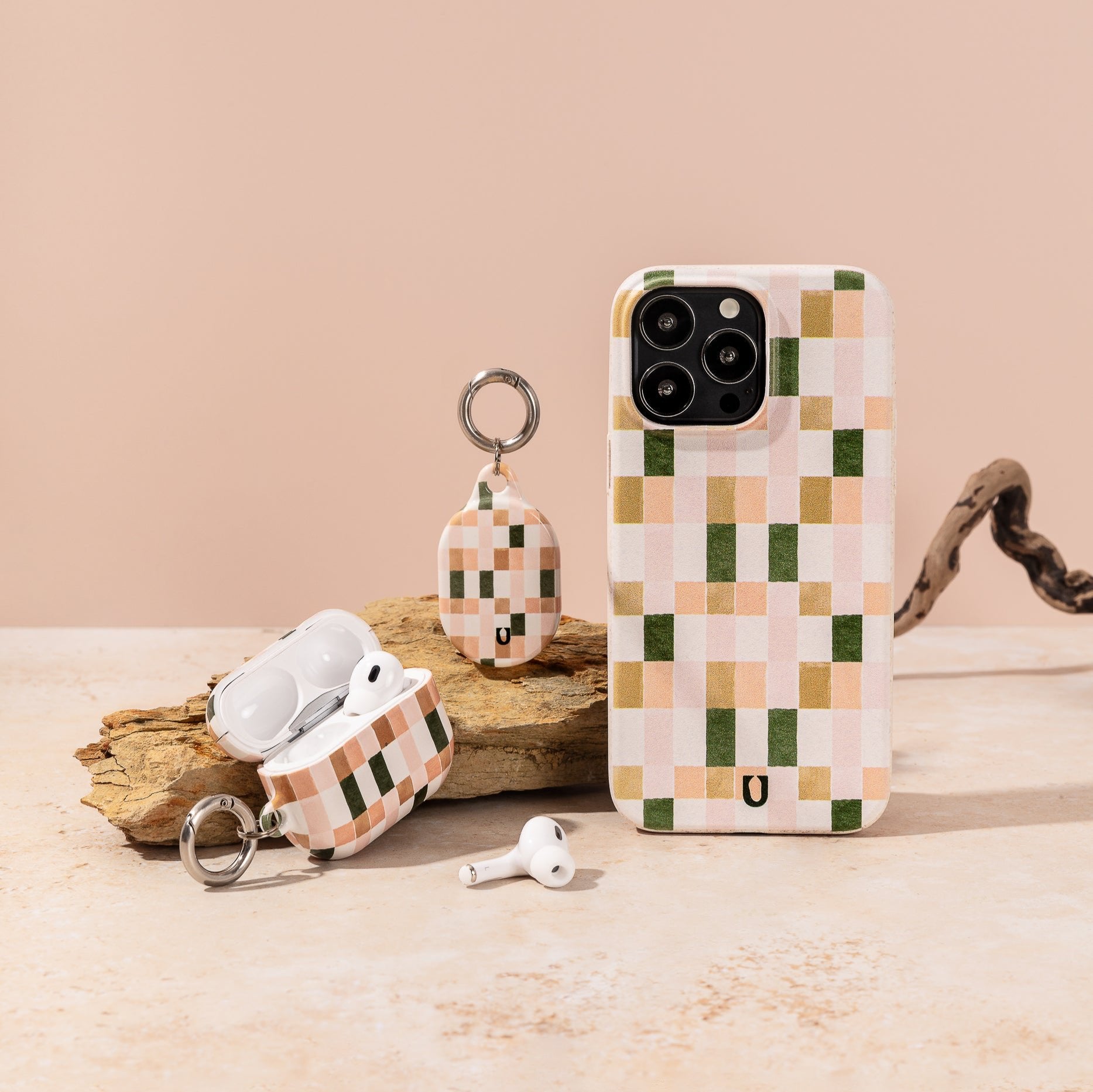 A Pink and Green Watercolour Eco-Friendly Phone Case, compatible with iPhone and Samsung Galaxy models, is displayed on a beige surface alongside matching AirPods case and AirTag holder with checkered patterns.