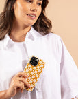 A woman in a white shirt holds a phone featuring the Star Terracotta Checker Eco-Friendly Phone Case, characterized by its yellow and white checkered design made from biodegradable materials, as she looks off to the side against a beige background.