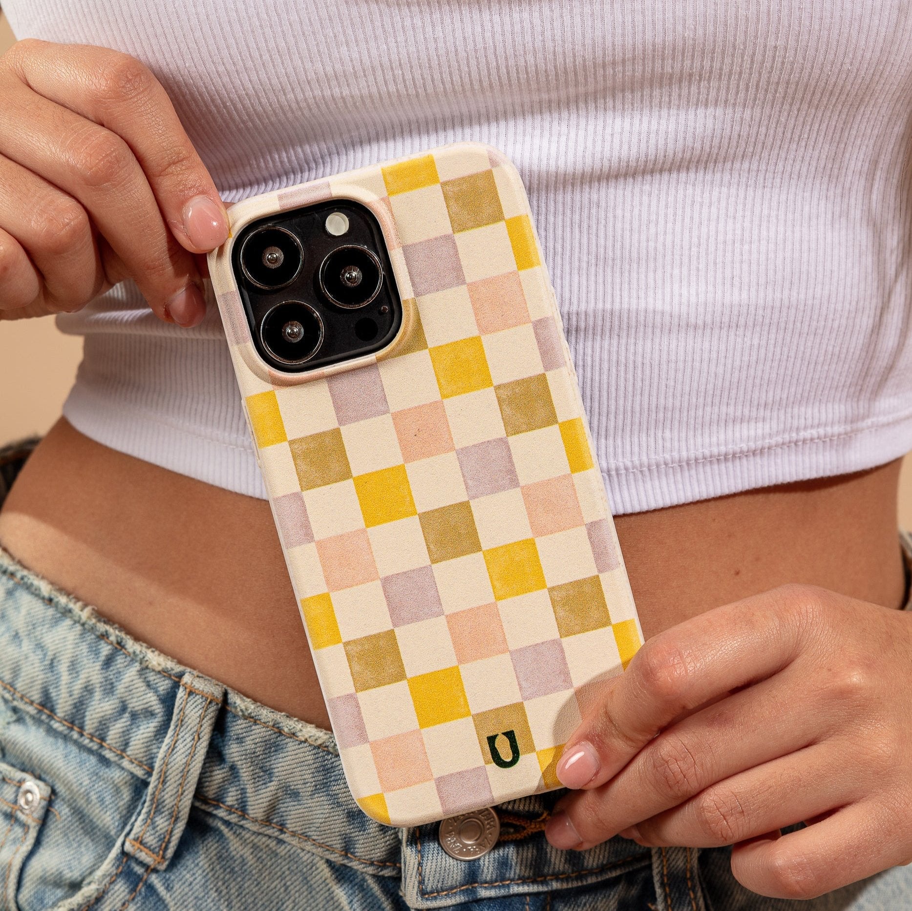 A person in a white crop top and denim jeans holds a phone with the Pink and Purple Checker Eco-Friendly Phone Case near their waist.