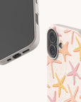 Two smartphones displayed with beige Starfish Eco-Friendly Biodegradable Phone Cases; one case features a pastel starfish pattern in pink, orange, and yellow. The phones are positioned to showcase both the front screens and the decorative case design.
