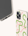 Two smartphones with beige Green and Pink Swirl Lines Eco-Friendly Phone Cases, each featuring playful green and pink squiggly designs. One phone displays the front screen angled upward, while the other reveals the back with camera lenses visible. These sustainable cases are compatible with both iPhone and Samsung Galaxy models, set against a light, neutral background.
