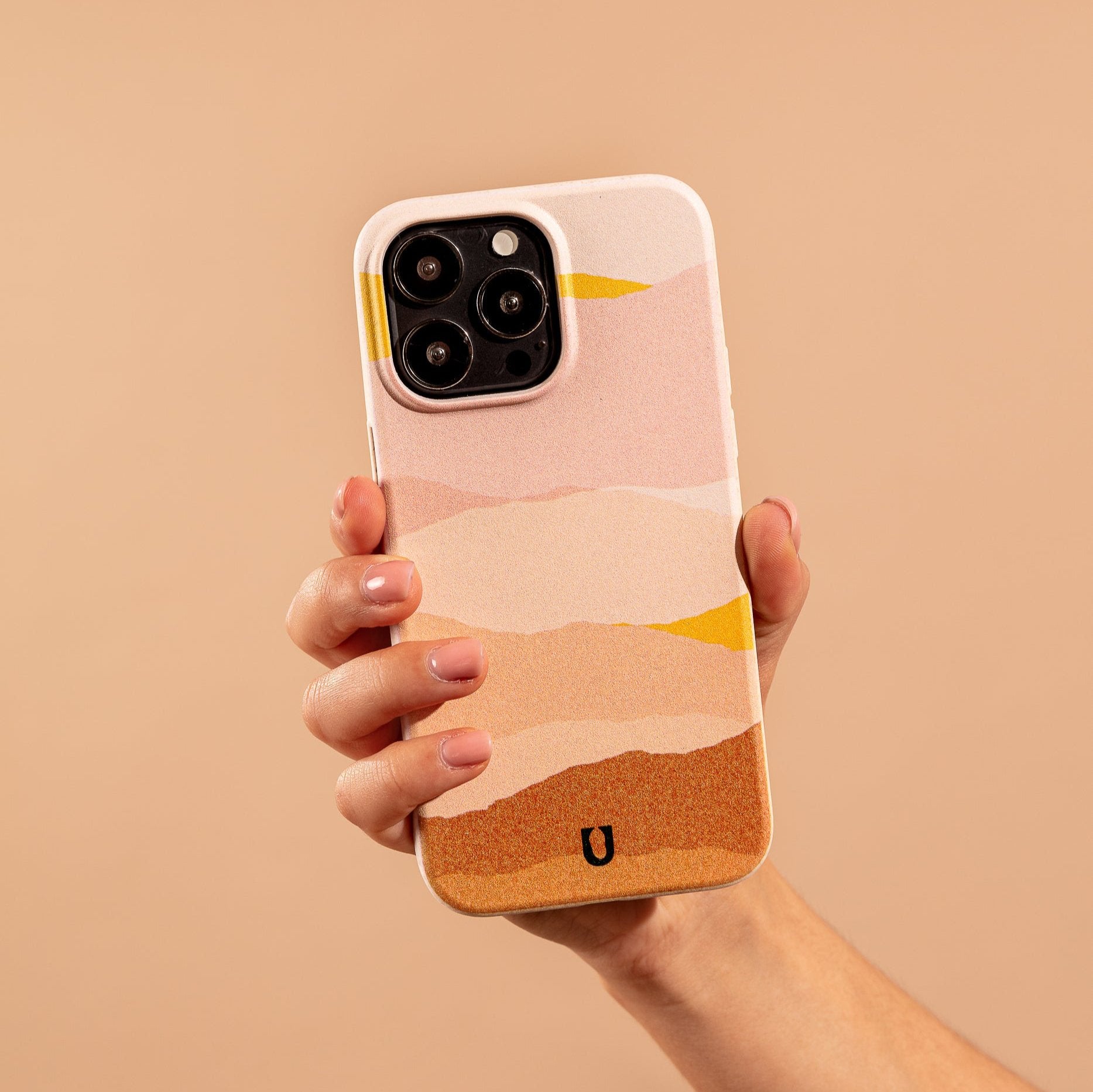 A hand holds the Mountains Eco-Friendly Phone Case, featuring an abstract, layered design in pink, peach, and yellow on beige. Compatible with iPhone and Samsung Galaxy models.