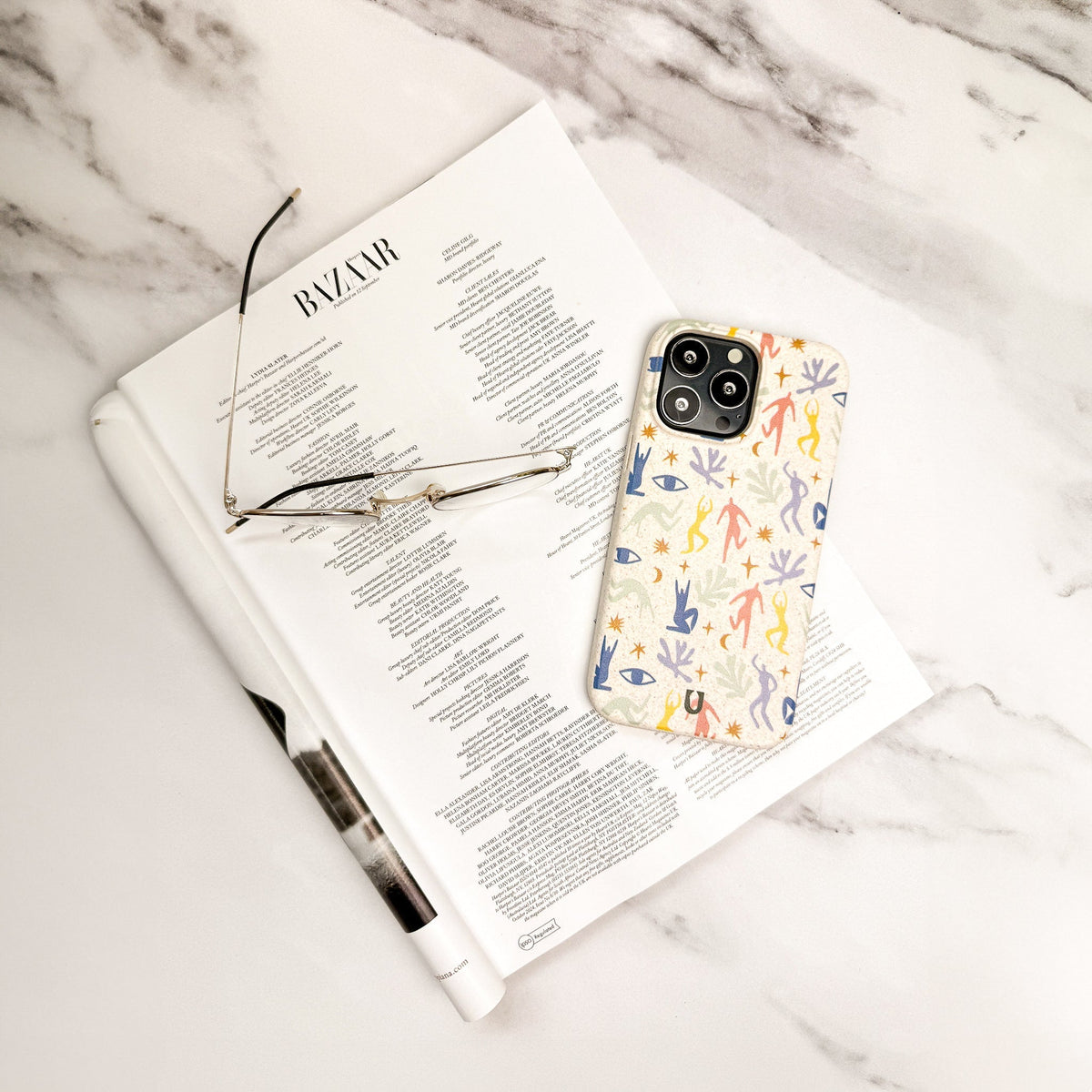 A folded magazine, a pair of eyeglasses, and a smartphone featuring the Joie de Vivre Eco-Friendly Phone Case are arranged on a white marble surface.