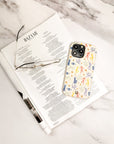 A folded magazine, a pair of eyeglasses, and a smartphone featuring the Joie de Vivre Eco-Friendly Phone Case are arranged on a white marble surface.