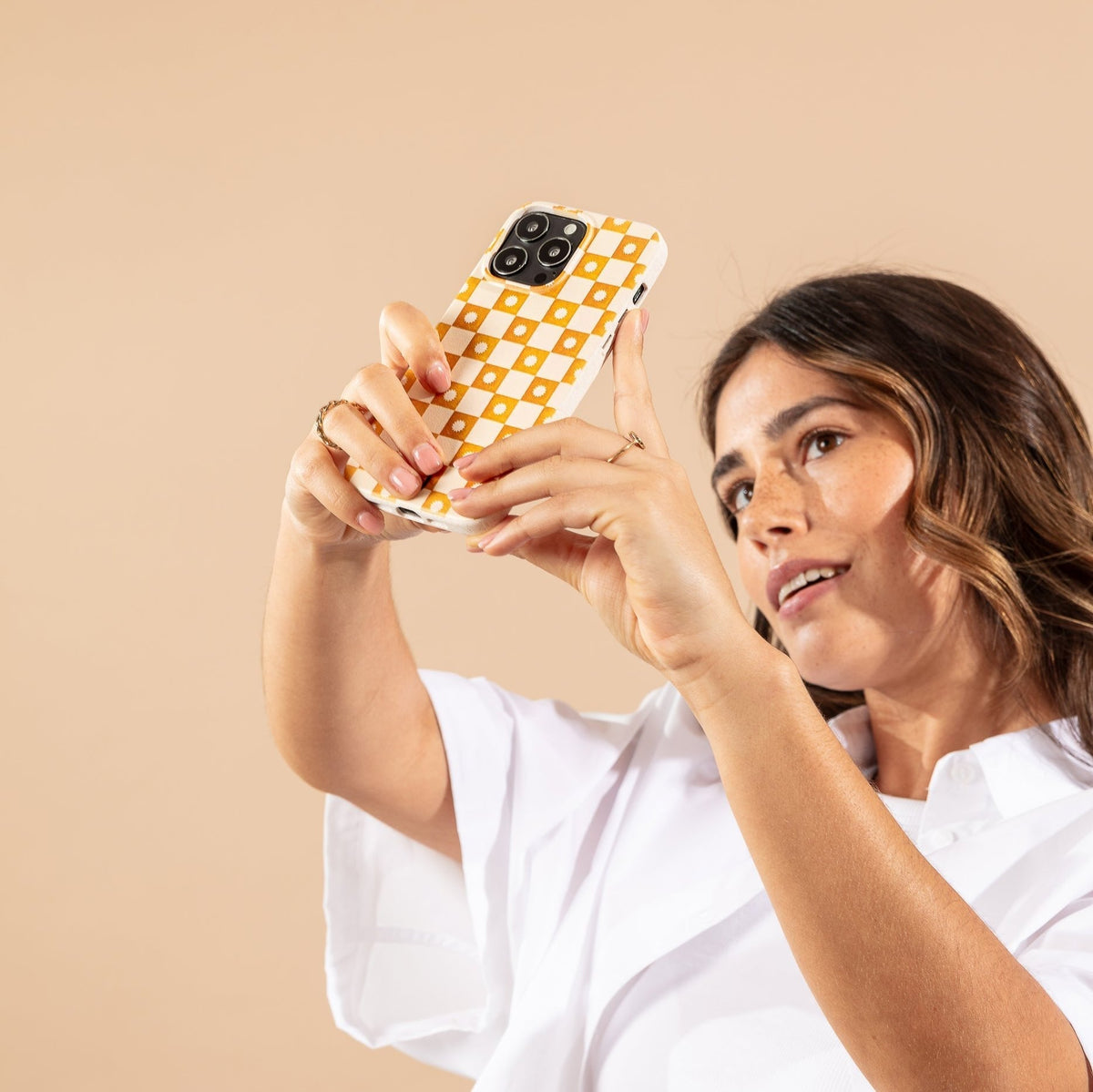 A woman in a white shirt holds up a phone with the Star Terracotta Checker Eco-Friendly Phone Case, featuring a yellow and white checkered design, appearing to take a selfie against a beige background.