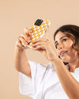 A woman in a white shirt holds up a phone with the Star Terracotta Checker Eco-Friendly Phone Case, featuring a yellow and white checkered design, appearing to take a selfie against a beige background.