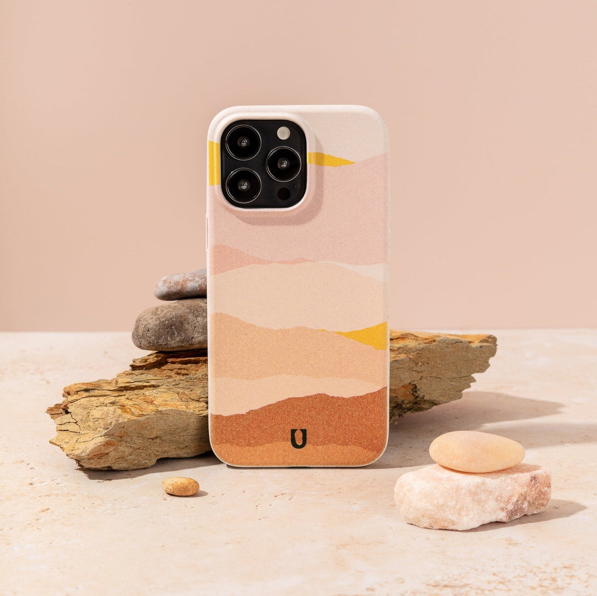 A smartphone with the Mountains Eco-Friendly Phone Case, featuring a pastel abstract design, leans against stacked stones and a beige rock amidst smooth pebbles on a light surface set against a muted peach background. This sustainable phone case is compatible with both iPhone and Samsung models.
