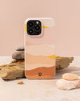 A smartphone with the Mountains Eco-Friendly Phone Case, featuring a pastel abstract design, leans against stacked stones and a beige rock amidst smooth pebbles on a light surface set against a muted peach background. This sustainable phone case is compatible with both iPhone and Samsung models.