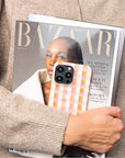 A person in a beige sweater holds a Harper’s Bazaar magazine and a tablet, with a Pink and Brown Gingham Eco-Friendly Phone Case visible in their hand.