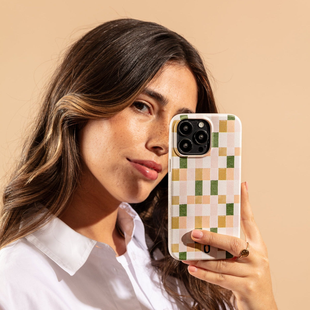 A woman with long brown hair holds a smartphone with a Pink and Green Watercolour Eco-Friendly Phone Case next to her face, looking at the camera. She wears a white shirt and a gold ring, against a beige background.