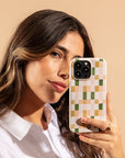 A woman with long brown hair holds a smartphone with a Pink and Green Watercolour Eco-Friendly Phone Case next to her face, looking at the camera. She wears a white shirt and a gold ring, against a beige background.