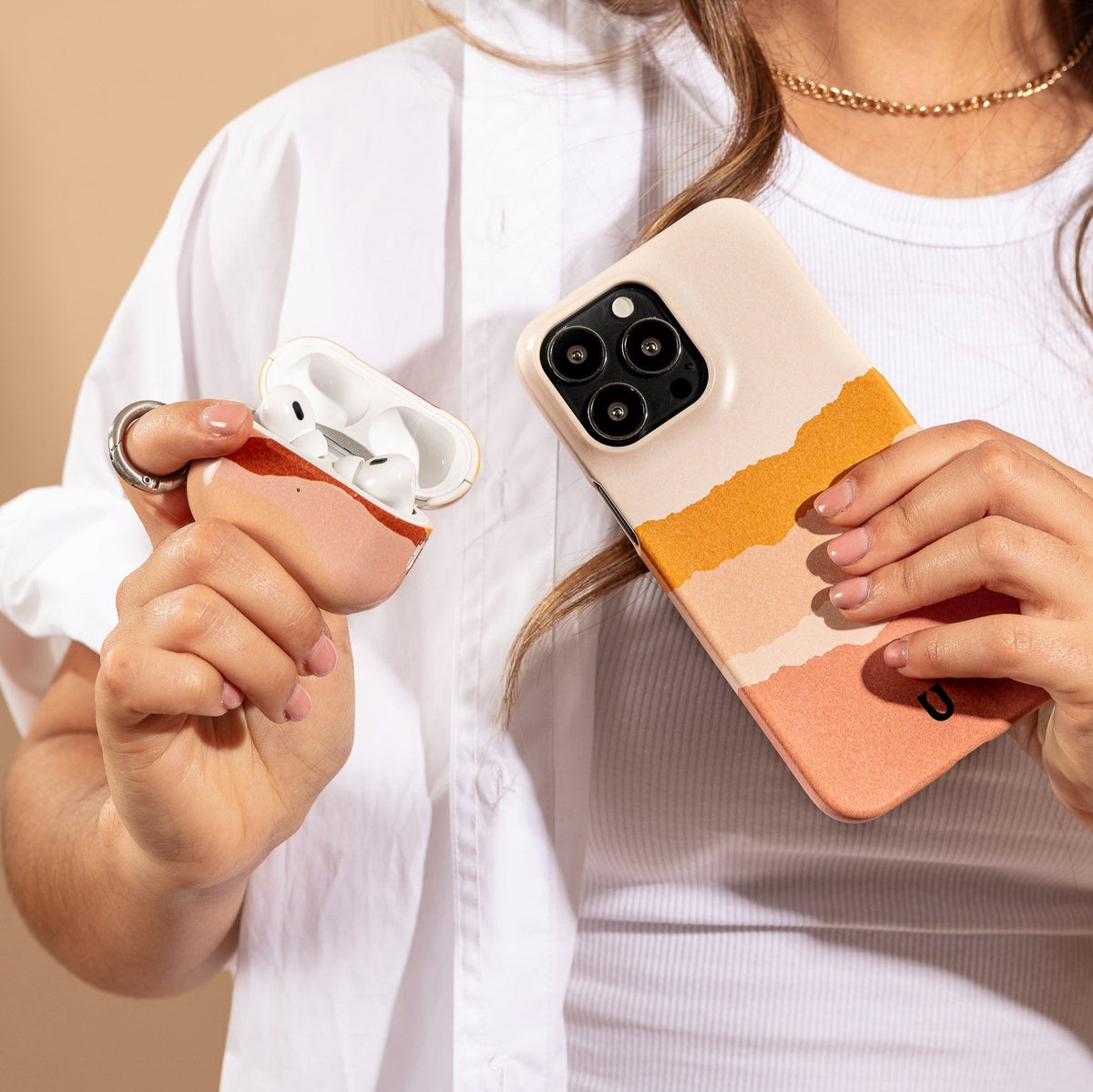 A person holds a smartphone encased in the Mountains Eco-Friendly Phone Case, featuring colorful stripes, in one hand and an open AirPods case in the other. The person is dressed in a white shirt with a gold necklace and rings. This sustainable phone case offers compatibility with both iPhone and Samsung devices, combining style and versatility.