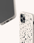 Two smartphones in Black Terrazzo Eco-Friendly Phone Cases—speckled off-white with black spots—are shown from different angles against a light background, showcasing the camera modules and the unique case texture.