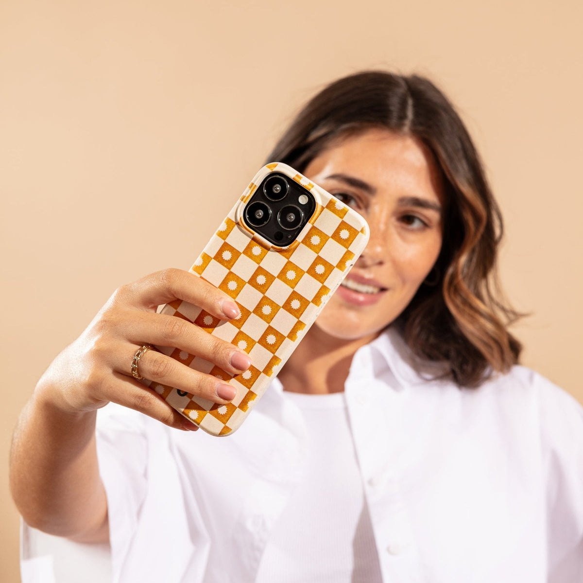 A woman in a white shirt takes a selfie against a beige background, holding up her smartphone fitted with the Star Terracotta Checker Eco-Friendly Phone Case featuring a yellow and white checkered pattern.