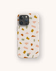 A smartphone with the Mini Confetti Eco-Friendly Phone Case, featuring a terrazzo pattern of scattered pastel geometric shapes in green, yellow, pink, and orange on a cream background. A small green U logo is positioned near the bottom center of this compostable case.