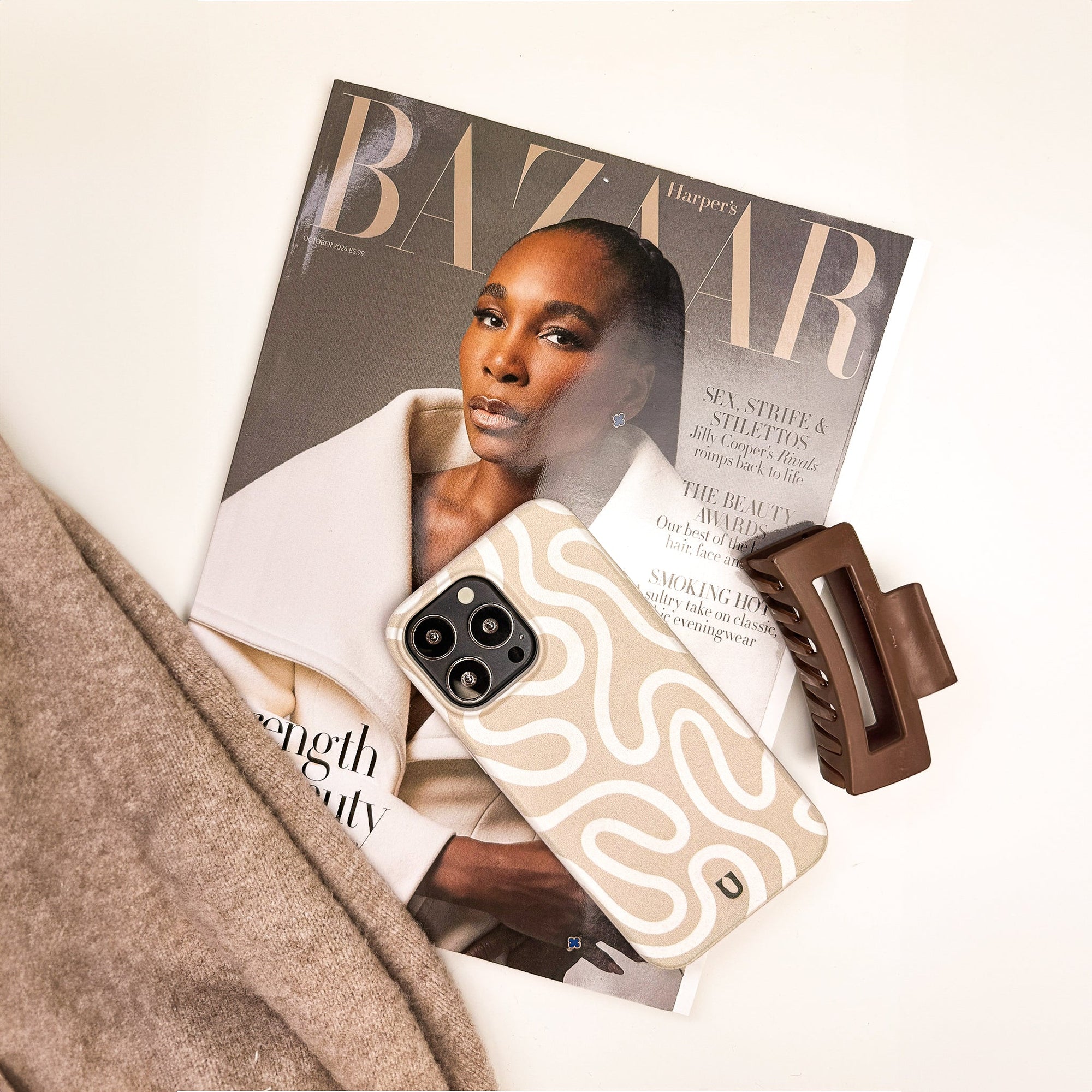 A Harper’s Bazaar magazine with a woman on the cover lies on a white surface, partially covered by a beige sweater. On top are a Neutral Swirl Lines Eco-Friendly Phone Case and a brown hair clip, creating a minimalist look.