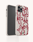 A smartphone sporting the Burgundy Bow Ribbons Eco-Friendly Phone Case, adorned with red bow designs on a white, subtly speckled background. A small green logo near the bottom of the case showcases its eco-friendly nature, and the phone features three rear camera lenses.