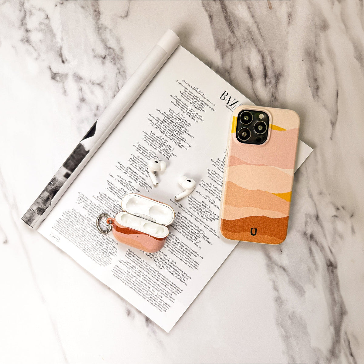 A smartphone with the Mountains Eco-Friendly Phone Case, wireless earbuds, and an earbud case are placed on an open magazine page on a white marble surface.