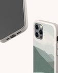 Two smartphones with beige speckled, biodegradable phone cases; one shows the screen and charging port, while the other displays the back, featuring the Blue Watercolour Landscape Eco-Friendly Phone Case design—a green abstract mountain motif and a triple camera setup.