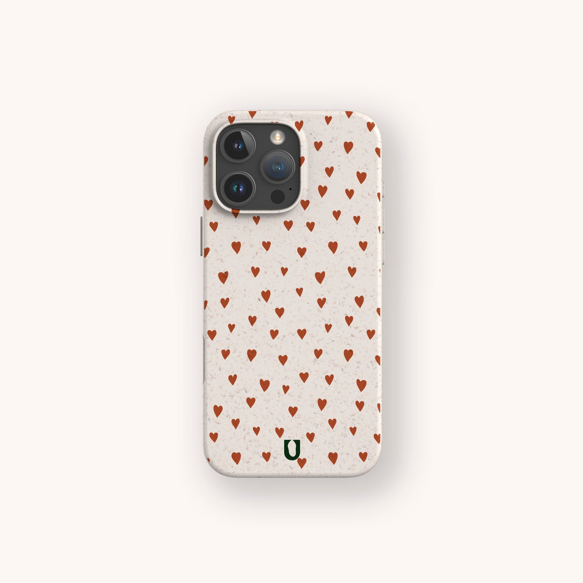 A smartphone with the Red Hearts Eco-Friendly Phone Case in beige, decorated with a pattern of small red hearts and a green horseshoe logo at the bottom. The phone’s camera features three lenses.