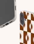 Two beige Sunseeker Eco-Friendly Biodegradable Phone Cases are featured: one showcases a brown and white checkerboard pattern with sunburst designs, while the other has a plain finish. Both cases have textured edges and precise openings for buttons and ports. Designed to be plastic-free, each Sunseeker case offers sustainable protection paired with stylish appeal.