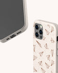 Two beige minimalist phone cases, both eco-friendly, are displayed against a plain light background. One case shows the phone’s screen and ports, while the other features a pattern of brown paper airplanes and is identified as the White Paper Planes Eco-Friendly Biodegradable Phone Case.