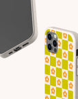 Two beige smartphone cases are displayed. One is the Green Flower Checker Eco-Friendly Phone Case, featuring a yellow and green checkerboard pattern with orange flowers, while the other shows the screen side and charging port of a phone. Both have a speckled, eco-friendly appearance and are available for iPhone and Samsung Galaxy models.
