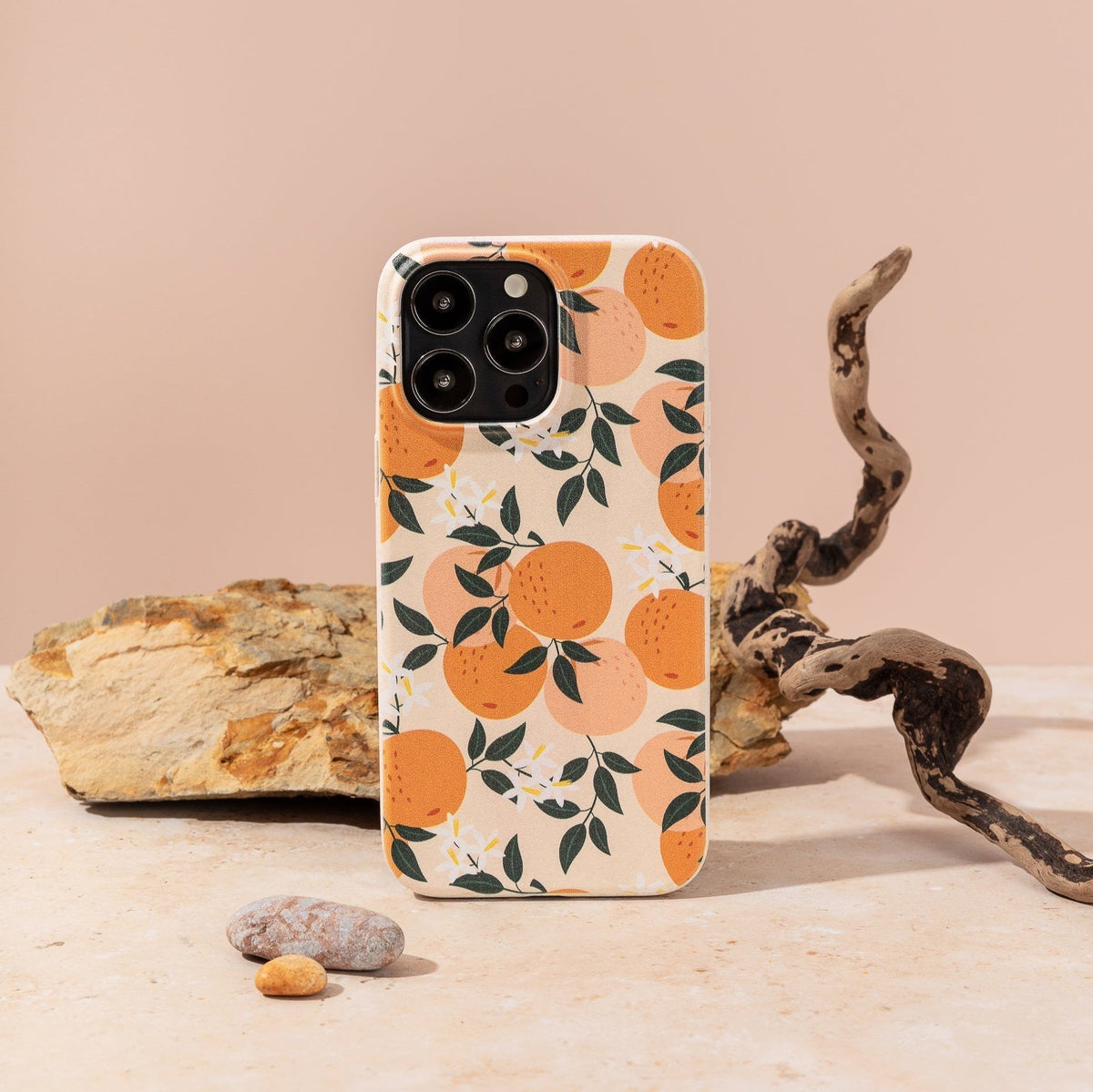 The Orange Blossom Eco-Friendly Phone Case, featuring an orange fruit and green leaves design, stands upright on a beige surface surrounded by rocks and a twisted branch against a neutral background.
