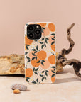 The Orange Blossom Eco-Friendly Phone Case, featuring an orange fruit and green leaves design, stands upright on a beige surface surrounded by rocks and a twisted branch against a neutral background.