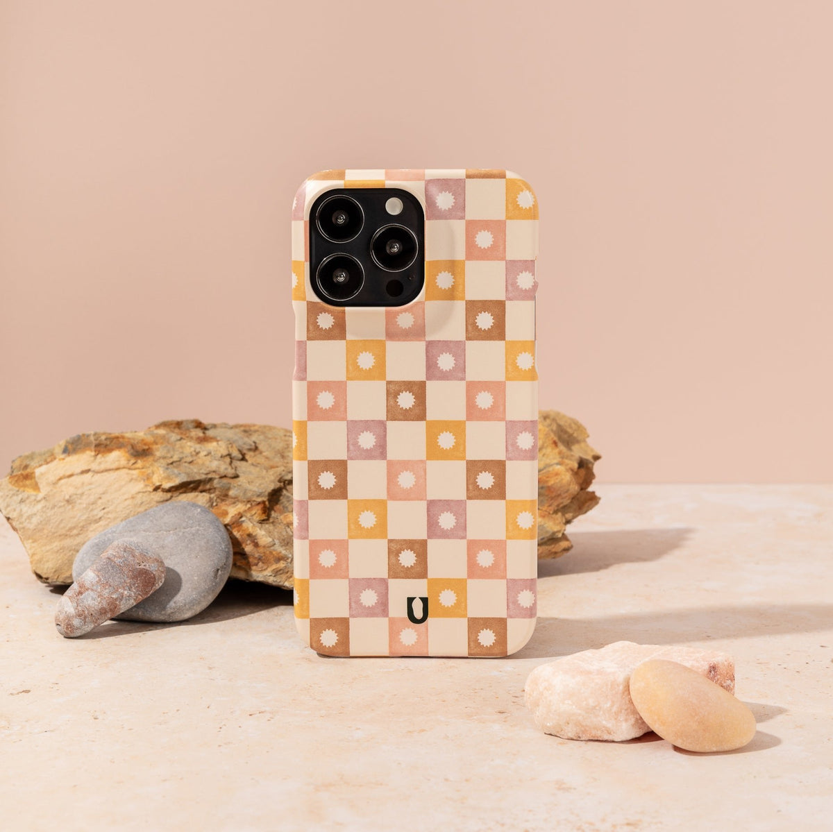 A smartphone stands upright on a stone surface with rocks in the background against a pale peach backdrop, featuring the Star Pink Checker Eco-Friendly Phone Case. This charming plant-based case showcases a checkered pattern in peach, pink, brown, and yellow adorned with small flowers and is compatible with both iPhone and Samsung Galaxy models.