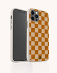 Brown Checker Eco-Friendly Phone Case shown on a smartphone, featuring a beige and orange checkerboard pattern with a small black and yellow logo at the bottom. The camera is clearly visible, while the case’s speckled biodegradable texture lends unique character and helps reduce environmental impact.