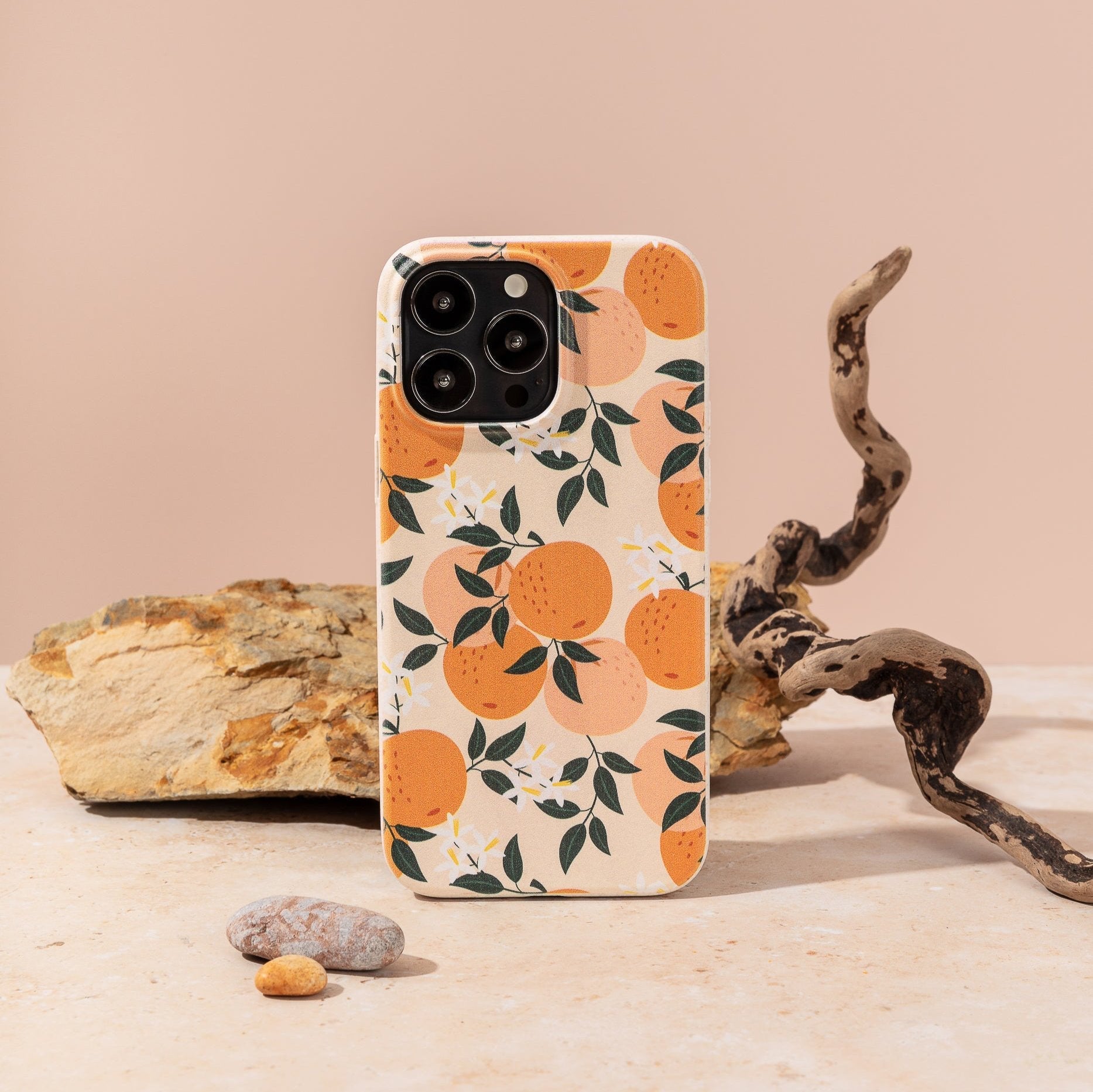 The Orange Blossom Eco-Friendly Phone Case stands upright on a beige surface, with a rock, twisted branch, and small pebbles in the background.