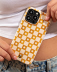 A person wearing a white ribbed crop top and blue jeans holds a phone with the Star Terracotta Checker Eco-Friendly Phone Case, which features a yellow and white checkered pattern adorned with small daisy designs.