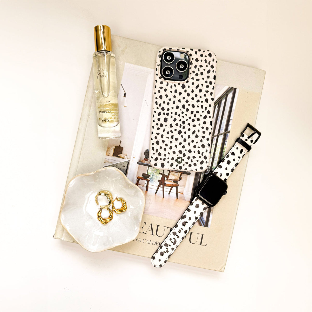 A flat lay of a book, a perfume bottle, a small dish with gold rings, and a watch is arranged on a white surface alongside the Dark Spots Eco-Friendly Phone Case for iPhone and Samsung Galaxy, featuring a minimalist black and white spotted design.