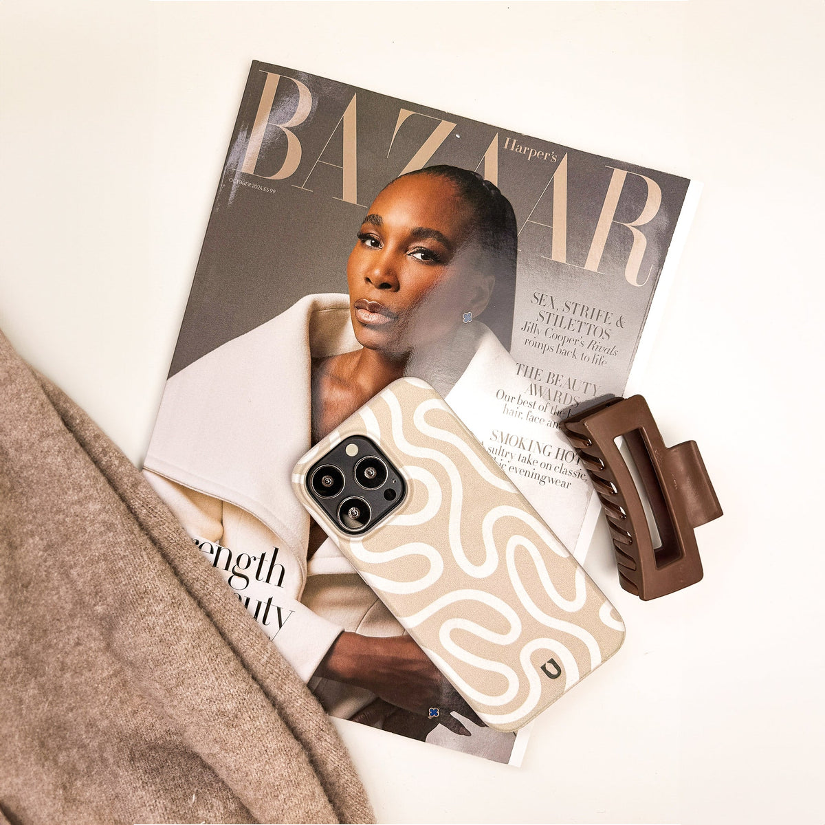 A Harpers Bazaar magazine featuring a fashion model on the cover, the Neutral Swirl Lines Eco-Friendly Phone Case—a beige minimalist case with a wavy pattern designed for iPhone and Samsung Galaxy users—a brown hair clip, and part of a tan sweater, all thoughtfully arranged on a white surface.
