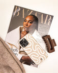 A Harpers Bazaar magazine featuring a fashion model on the cover, the Neutral Swirl Lines Eco-Friendly Phone Case—a beige minimalist case with a wavy pattern designed for iPhone and Samsung Galaxy users—a brown hair clip, and part of a tan sweater, all thoughtfully arranged on a white surface.