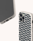 Two smartphones with off-white cases; one case is the Black Flower Checker Eco-Friendly Phone Case, which features a checkerboard design with black and white flowers, while the other phone displays its screen and port details. Both are set against a plain, light background.