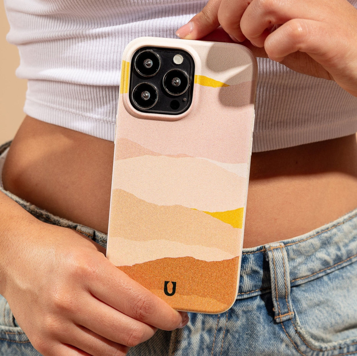 A person in a white crop top and blue jeans holds a phone with the Mountains Eco-Friendly Phone Case—a sustainable accessory compatible with iPhone and Samsung devices. The pastel-colored, abstract-patterned case features visible camera lenses and showcases shades of pink, orange, yellow, and beige.
