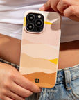 A person in a white crop top and blue jeans holds a phone with the Mountains Eco-Friendly Phone Case—a sustainable accessory compatible with iPhone and Samsung devices. The pastel-colored, abstract-patterned case features visible camera lenses and showcases shades of pink, orange, yellow, and beige.